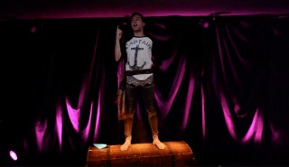 (Image: Donovan Stands on a Sea Chest with Curtains Behind
  Illuminated in Slashes of Purple Light)