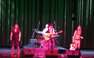 (Image: The Band Lighted in Pink in front of a Pleated Curtain
 Covered with White Slashes)