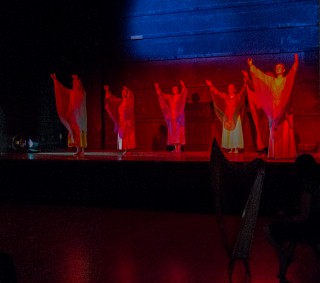 (Image: A Harpist in Silhouette watches Dancers
  in Red with their Arms Raised)
