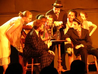 (Image: The Cast Crowds Around a Table in the Speakeasy)